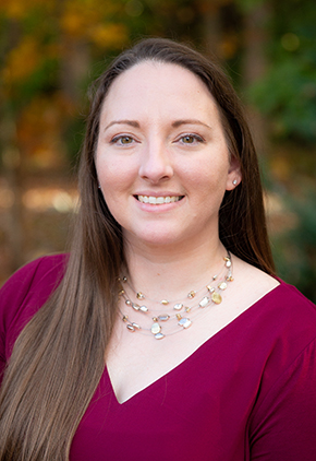 Lexi Dickens smiles outside for a professional headshot on a fall day. She is wearing a deep red top and three strand necklace.