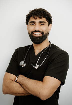 Dr. Vazul Munjal smiles for a professional headshot in front of a white wall. He is wearing black scrubs and has his stethoscope around his neck with his arms crossed in front of his chest.