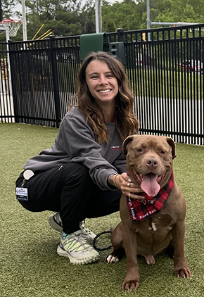 Jen McConnell squats outside next to a brown pit bull. She's smiling, wearing black joggers and a grey quarter zip sweatshirt.