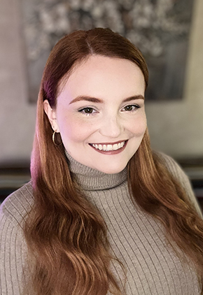 Brooke Mckeand smiling in front of a tan wall with a large abstract art piece. She is wearing a taupe turtleneck.