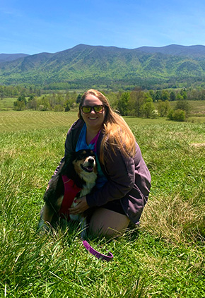 A woman kneels in a meadow of grass while hugging a black and white dog.