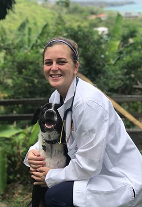 Dr. Brianna Jacobs smiles outside while kneeling next to, and hugging, a black and white dog. She is wearing her whitecoat and has a stethoscope around her neck.