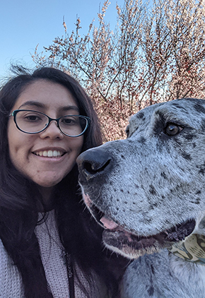 Andrea Hernandez smiles outside with one of her dogs. She is wearing a light pink sweatshirt and has black rounded glasses. Her dog is large, grey and has black spots.