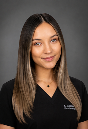 Dr. Katherine Holmes smiles for a professional headshot in front of a grey background. She is wearing black scrubs that feature her embroidered nail.