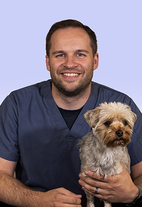 Dr. Dillon Scotts smiles in a studio for a professional photo. He is wearing navy scrubs with his arm around a small terrier who is standing on a table.