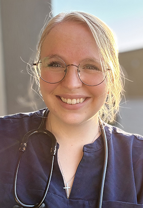 Dr. Sydney Elwood smiles for a photo outside. She is backlit by the sun and wearing navy scrubs with a black stethoscope around her neck.