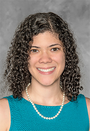 Dr. Victoria DeMello smiles for a professional headshot in front of a grey background. She is wearing a teal sleeveless top and a pearl necklace.