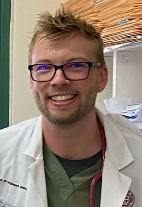 Dr. Zachary Jacobsen smiles inside a hospital office. He is wearing olive green scrubs and his white coat.