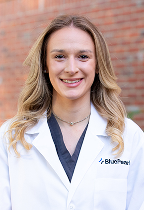 Dr. Leah Orton smiles outside in front of a brick building. She is wearing a slate blue blouse and her BluePearl whitecoat.