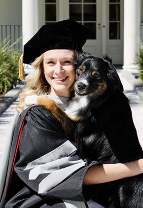 Dr. Leah Orton smiles outside of a building at Ross University. She is squatting next to her Australian Shepherd, Poppi. Poppi is standing on her hind legs while she and Dr. Orton hug. Dr. Orton is wearing her doctoral regalia.