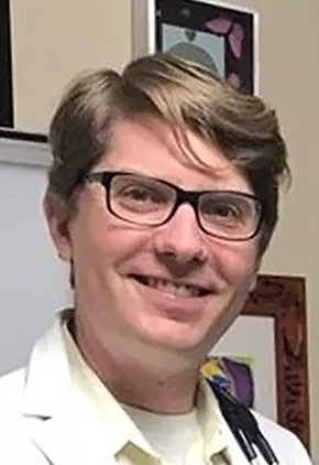 Dr. Tony Kahn smiles for a photo in front of framed photos. He is wearing a white collared shirt and his whitecoat.