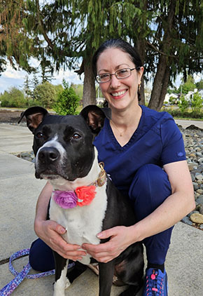Dr. Naomi Weinberg smiles and kneels outside next to one of her dogs. She is wearing navy blue scrubs and there are lush trees in the background.