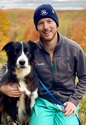 Dr. Chris Stretton kneels in a field with his border collie, Frank. Dr. Stretton is wearing teal scrubs and a grey softshell jacket.