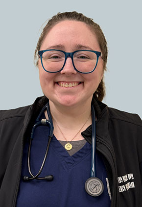 Dr. Cassidy McCarthy smiles for a professional headshot in front of a light blue-grey background. She is wearing navy scrubs and a black jacket with her stethoscope around her neck.
