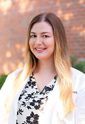 Dr. Damova Star smiles outside in front of a brick building. She is wearing a white blouse that features black flowers and her BluePearl whitecoat.