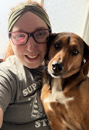 Dr. Liz Olson smiles while basking in the warmth of the sun coming from a living room window while her dog is in her lap.