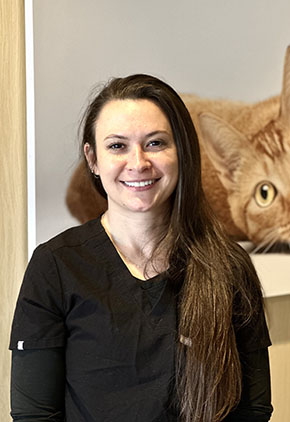 Dr. Valerie Cwiek smiles for a headshot in the lobby of BluePearl. A large photo of an orange cat laying down is hanging on the wall behind her. She is wearing black scrubs.