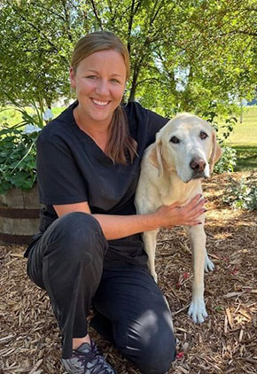 Dr. Danielle Renstrom smiles outside while kneeling next to her yellow lab. They are in the mulch area in front of a green tree. Dr. Renstrom is wearing navy scrubs.