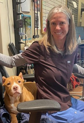 Dr. Kacee Lingner smiles while sitting in an office chair with a tan pit bull on the seat behind her. She is wearing blue scrub bottoms and a dark brown scrub top.