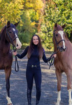 Dr. Jennifer Murphy smiles outside on a trail holding the reins of her horses, Petey and Baloo. She is in dark riding pants, a black turtleneck and black riding boots.