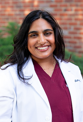 Dr. Vi Dharmasena smiles outside in front of a brick building. She is wearing maroon scrubs and her BluePearl whitecoat.