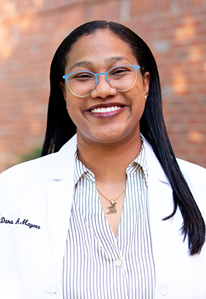Dr. Dara Mayers smiles outside in front of a brick building. She is wearing a light blue pinstripe blouse with her personalized whitecoat.