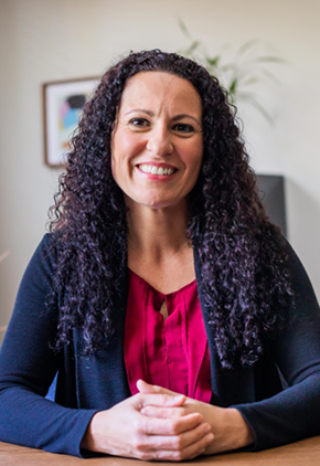 Dr. Carolina Medina sits at a desk in her office and smiles. She has her hands together and resting atop the table. She is wearing a pink blouse and navy cardigan. The background features the leaves of a plant behind her head and a framed abstract art piece hanging on the light tan wall.