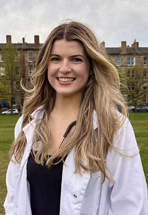 Dr. Chloe Williams smiles outside on the lawn at the University of Edinburgh, a campus building is featured in the background. She is wearing a black top and a white coat with a stethoscope around her neck.