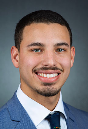 Dr. David Dominguez smiles for a professional headshot. He is wearing a dusty blue suit with a white dress shirt and dark navy tie.