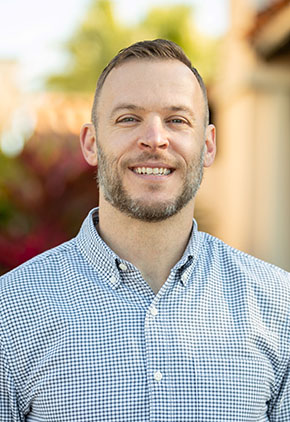 Dr. Ben O'Kelley smiles outside for a professional headshot. He is wearing a light blue checkered button-up shirt.