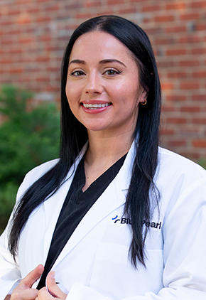 Dr. Paige Christiana smiles for a professional headshot outside in front of a brick building. She is wearing black scrubs and her BluePearl whitecoat.