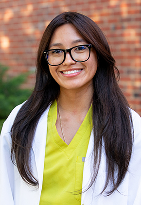 Dr. Brianna Espinosa smiles outside in front of a brick building for a professional headshot. She is wearing neon yellow scrubs and her BluePearl whitecoat.