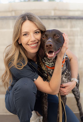 Dr. Danielle Perkon smiles outside while kneeling next to, and wrapping her arms around, one of her dogs in front of a fountain. She is wearing grey scrubs.