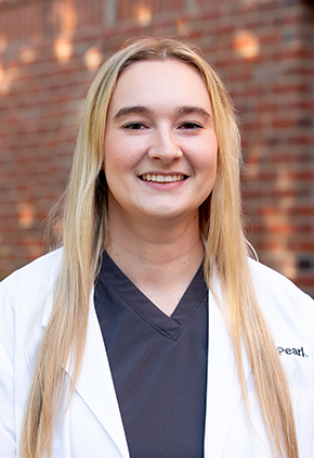Dr. Stephanie Steinke smiles outside in front of a brick building. She is wearing navy scrubs and her BluePearl whitecoat.