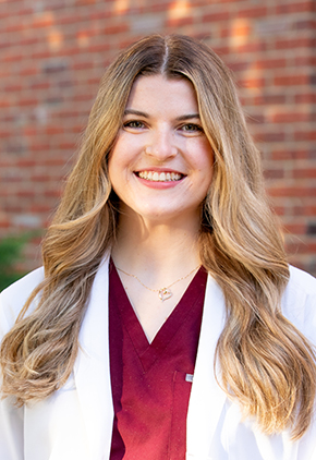 Dr. Chloe Williams smiles outside in front of a brick building. She is wearing red scrubs and her BluePearl whitecoat.