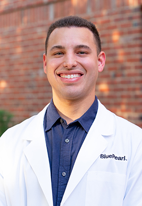 Dr. David Dominguez smiles outside in front of a brick building. He is wearing a blue dress shirt and his BluePearl whitecoat.