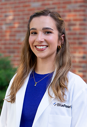 Dr. Samantha Forjan smiles outside in front of a brick building. she is wearing a blue blouse and her BluePearl whitecoat.