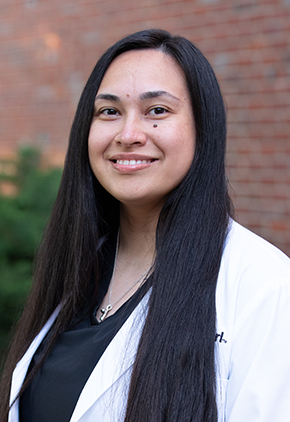 Dr. Leslie Cajas Chon smiles outside in front of a brick building. She is wearing black scrubs and her BluePearl whitecoat.