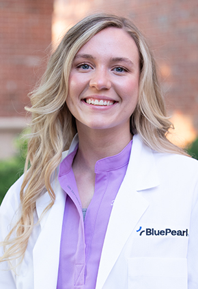Dr. Kate Williams smiles outside in front of a brick building. She is wearing a lavender blouse and her BluePearl whitecoat.