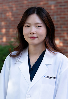 Dr. Ella Lee smiles outside in front of a brick building. She is wearing black scrubs and her BluePearl whitecoat.