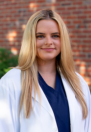 Dr. Cayla Morris smiles outside in front of a brick building. She is wearing navy scrubs and her BluePearl whitecoat.