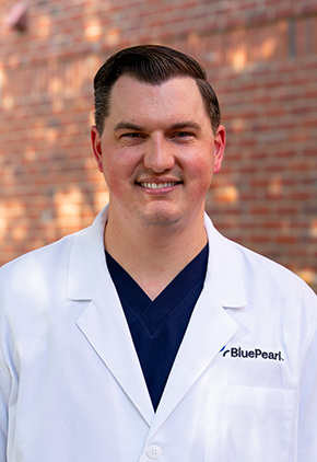 Dr. Patrick Dittmer smiles for a professional headshot outside in front of a brick building. He is wearing black scrubs and his BluePearl whitecoat.