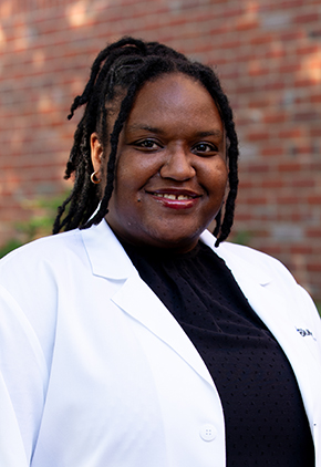 Dr. Mya Green smiles outside in front of a brick building. She is wearing a black turtleneck and her BluePearl whitecoat.