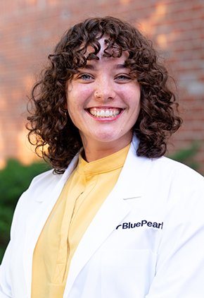 Dr. Savannah Bowles smiles outside in front of a brick building. She is wearing a yellow blouse and her BluePearl whitecoat.