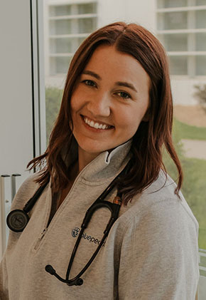 Dr. Kaitlynn Solberg smiles for a headshot next to a large window that opens to a courtyard. She is wearing a grey BluePearl quarter-zip and has a stethoscope around her neck.