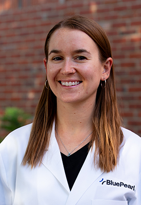 Dr. Alice Katz smiles outside for a professional headshot in front of a brick building. She is wearing black scrubs and her BluePearl whitecoat.