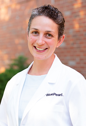 Dr. Rebekah Swartz smiles outside in front of a brick building. She is wearing a pale blue blouse and her BluePearl whitecoat.