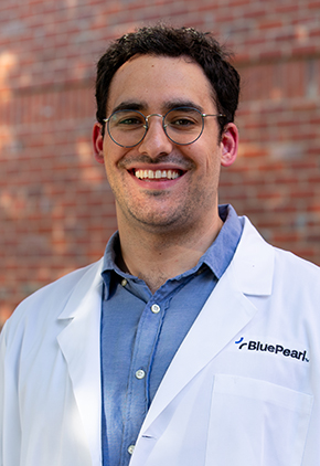 Dr. Alexander Petruso smiles outside in front of a brick building. He is wearing a blue dress shirt and his BluePearl whitecoat.