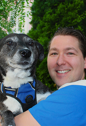 Dr. Marko Palumbo smiles outside while holding his black and white terrier mix, Quartz, in his arms. He is wearing a blue shirt.