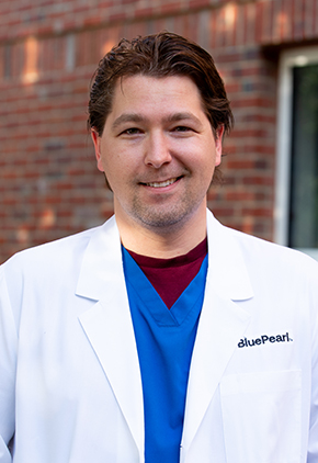 Dr. Marko Palumbo smiles outside in front of a brick building. He is wearing blue scrubs and his BluePearl whitecoat.